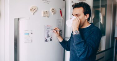 young man writing reminder on fridge and drinking coffee at home