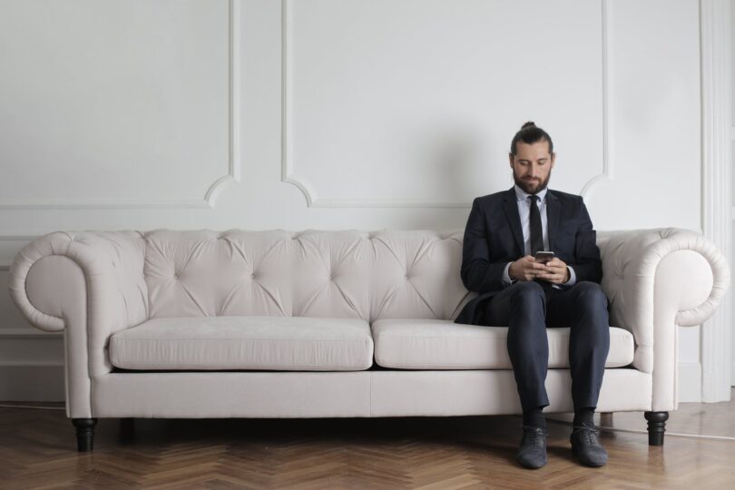 man sitting on white couch using smartphone