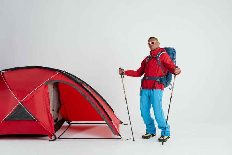 a man in red jacket and blue pants holding a trekking poles while standing near the red tent